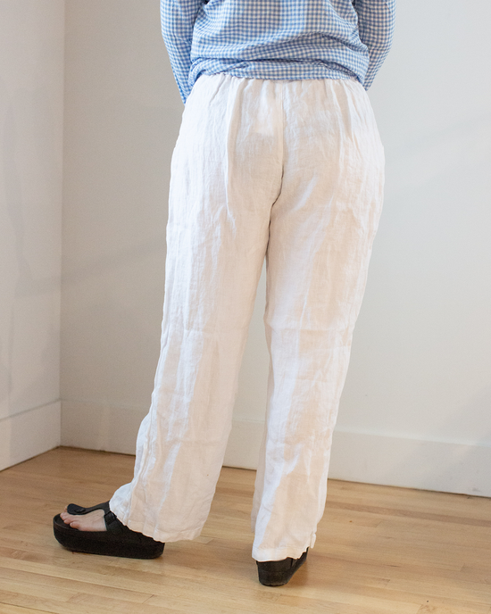 A person standing against a wall wearing CP Shades' Tess Pant HW Linen Twill in White trousers and black sandals.