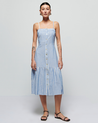 Woman in a Nation LTD Luciana Stripe Single Tier Dress in Parisian Blue, standing against a plain background. She has tattoos and is wearing black sandals.