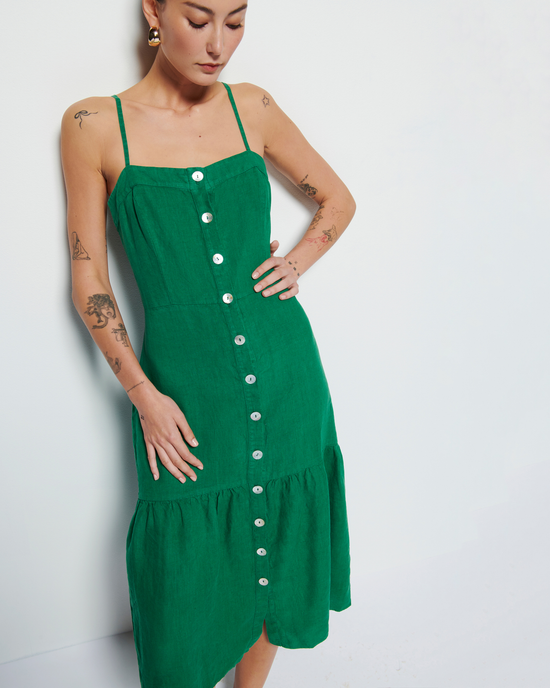 A woman in a Luciana Single Tier Dress in Verdant Green by Nation LTD with white buttons stands against a white background, her hand resting on her hip, showcasing tattoos on her arms.