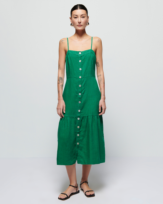 A woman stands facing the camera, wearing a Nation LTD Luciana Single Tier Dress in Verdant Green with button details and black sandals, set against a plain light background.