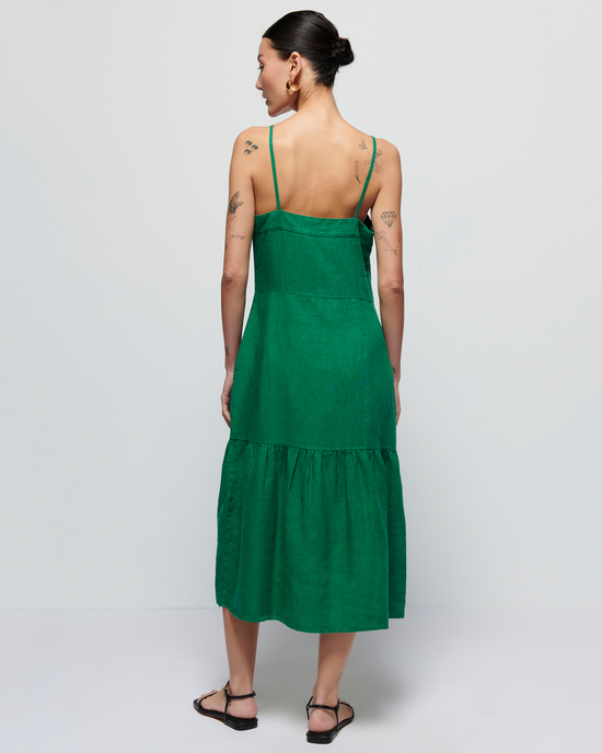 A woman standing with her back to the camera wearing a Luciana Single Tier Dress in Verdant Green by Nation LTD with thin straps and tattoos visible on her upper arms.