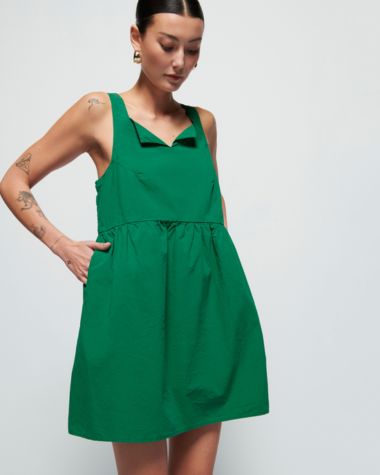 Woman in Nation LTD's Solie Dress in Verdant Green with shirring around the waist, posing against a light background, with visible tattoos on her left arm.