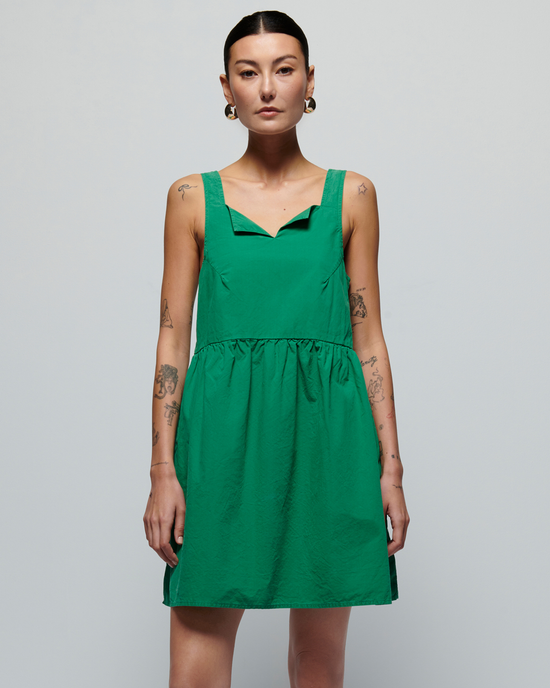 A woman with tattoos wearing a Nation LTD Solie Dress in Verdant Green featuring shirring around the waist stands against a plain light gray background.