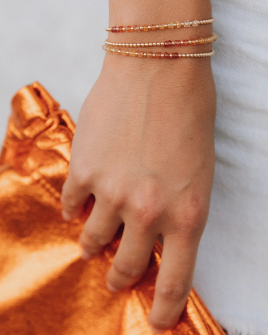 A person's wrist adorned with delicate beaded bracelets, featuring the 2MM Sig Bracelet with Sunrise Ombre & Yellow Gold by Karen Lazar Design, against a gold fabric background.