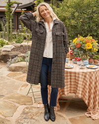 A woman smiling outdoors, wearing a Frank & Eileen Gavin Italian Wool Shirt Jacket in Black & Blue Textured Plaid, jeans, and boots, standing near a table set with flowers and food.