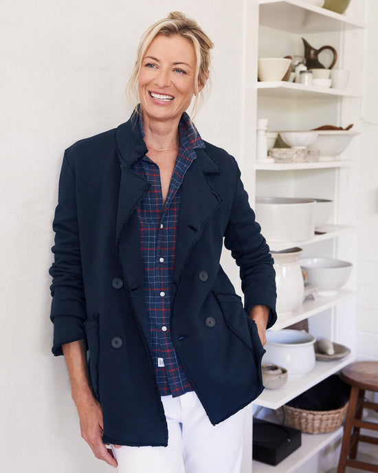 A smiling blonde woman wears a Frank & Eileen Belfast Sweatshirt Peacoat in British Royal Navy over a plaid shirt, standing in a kitchen with open shelves holding white dishes and bowls.