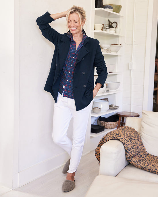 A smiling woman leans against a white wall indoors, wearing the Frank & Eileen Belfast Sweatshirt Peacoat in British Royal Navy over a plaid shirt, paired with white pants and gray slippers. Shelves and a white couch are visible in the background.