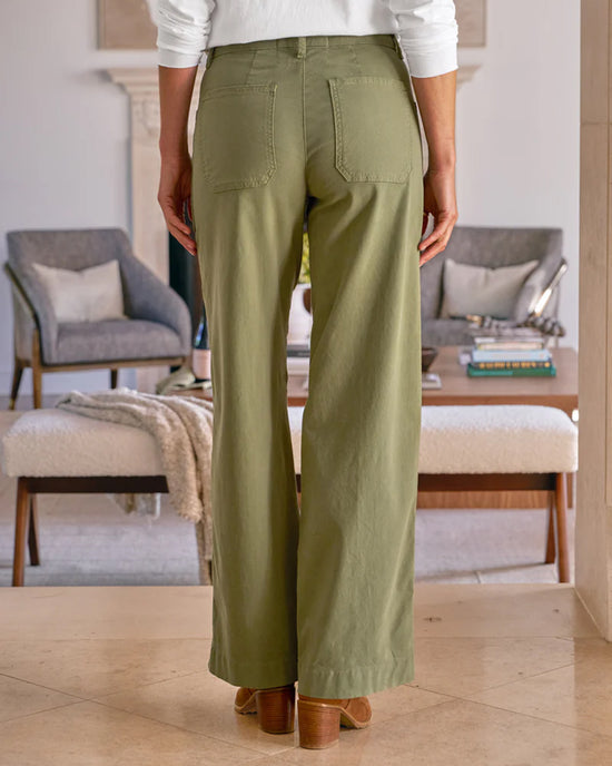 A person stands indoors facing away, wearing Frank & Eileen's Limerick Wrap Pocket Pant in Olive with light brown heeled shoes, set against a modern, neutral-toned living room with contemporary chairs and a coffee table.