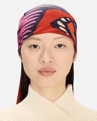 A person with straight dark hair wears the Inoui Editions Square / Carre 130 Papillons in Red as a vibrant headscarf and a cream ribbed sweater, looking neutrally at the camera against a plain light background.