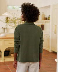 A person with curly hair, wearing Frank & Eileen’s Patrick Popover Henley in Caper Terry and white pants, stands with their back to the camera in a bright, modern kitchen.