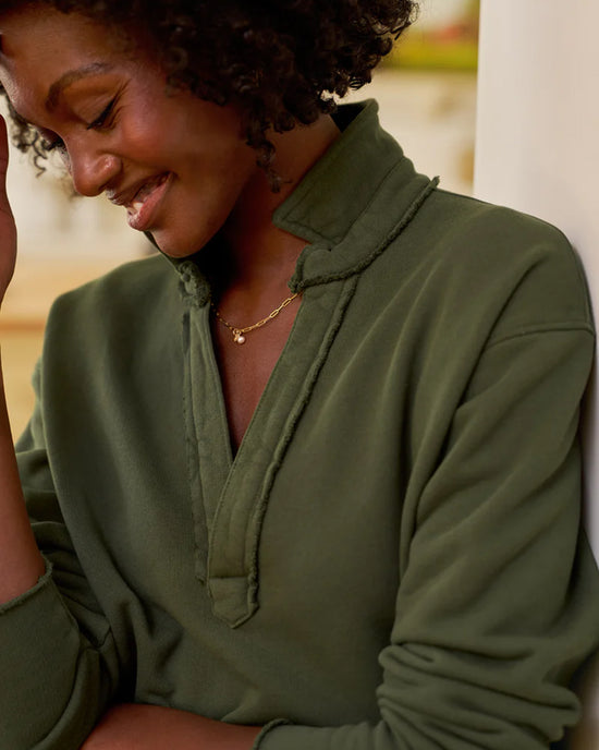 A woman with curly hair smiles gently, looking down. She wears a Frank & Eileen Patrick Popover Henley in Caper Terry and a delicate gold necklace, with a softly blurred background.