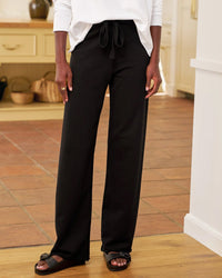 A person stands indoors in Frank & Eileen Catherine Long Sweatpant in Black, paired with a white long-sleeve top and black slide sandals. The kitchen background includes baskets and wooden cabinets.
