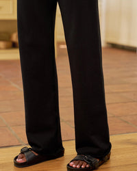 A person standing indoors wearing Frank & Eileen Catherine Long Sweatpant in Black and black buckle sandals, with brown tile flooring and wooden shelves in the background.