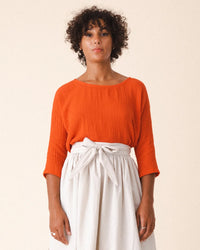 A person with curly hair wears Prairie Underground’s Third Wave in Electric Red—a boxy, long-sleeve top—paired with a high-waisted, light skirt with a tied belt, standing against a plain background.