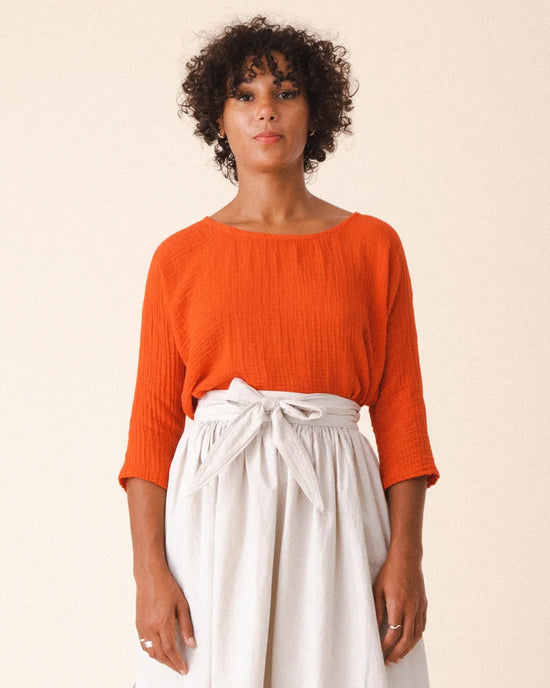 A person with curly hair wears Prairie Underground’s Third Wave in Electric Red—a boxy, long-sleeve top—paired with a high-waisted, light skirt with a tied belt, standing against a plain background.