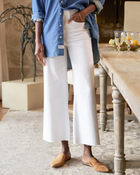 A person wearing a blue Italian Dream Denim shirt and Frank & Eileen's Galway Gaucho in White Denim stands beside a wooden table, with one hand on their hip and brown loafers on their feet.