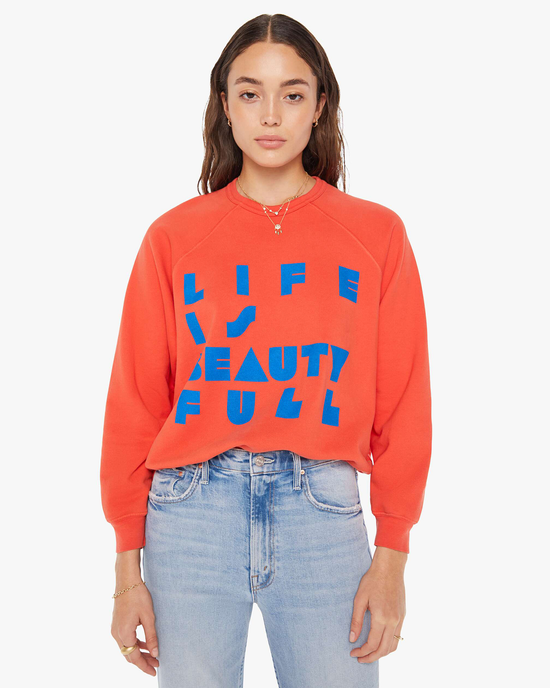 Woman in a slightly cropped silhouette, orange Mother pullover with raglan sleeves and The Biggie Concert in Live is Beauty Full phrase in blue letters.