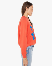 Side profile of a woman wearing an orange Mother The Biggie Concert in Live is Beauty Full pullover with blue text and blue jeans, featuring a slightly cropped silhouette.