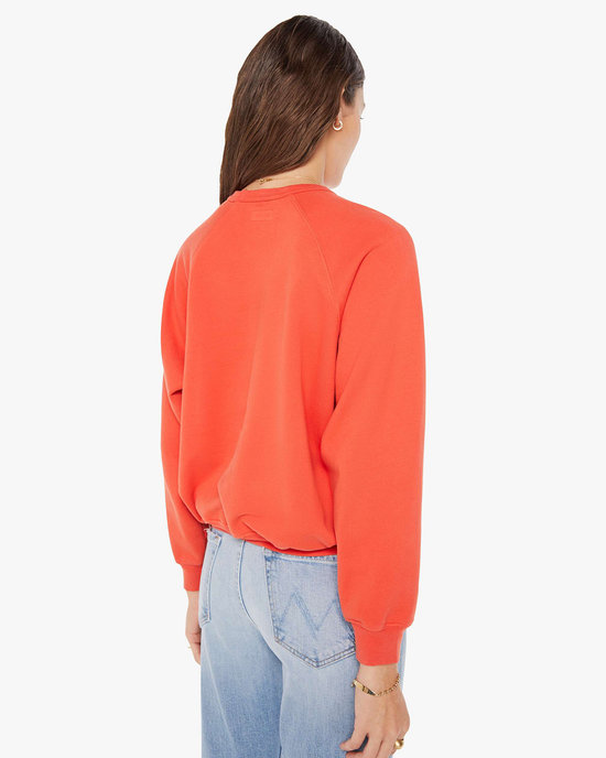 Woman standing with her back to the camera wearing an orange Mother pullover with a raglan sleeve and blue jeans.