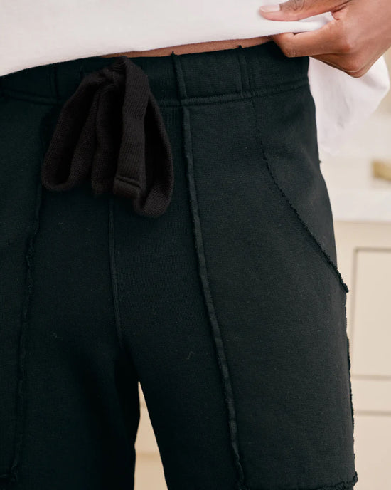 Close-up of a person wearing Frank & Eileen Penny Patch Pocket Sweatpant in Black and a white shirt, with one hand holding up the waistband to reveal the drawstring.