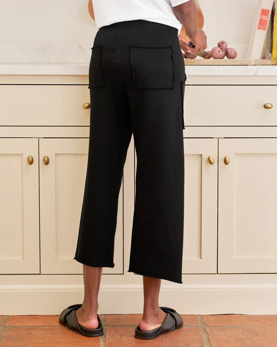 A person stands in a kitchen wearing Frank & Eileen’s Penny Patch Pocket Sweatpant in Black, black slide sandals, and a white shirt as they hold potatoes on the counter. The image is taken from behind.