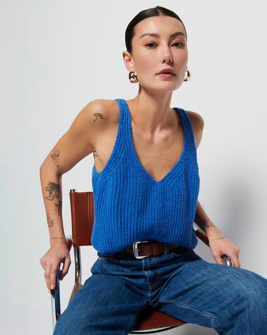 A woman with a tattoo on her arm seated in a chair, wearing a Nation LTD Cece Sweater Tank in Palace Blue, jeans, and large earrings, against a plain background.
