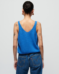 Rear view of a woman with tattoos wearing a Nation LTD Cece Sweater Tank in Palace Blue and denim jeans, standing against a plain background.