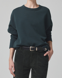 A person wearing an oversized dark green Subert Sam sweatshirt and black Citizens of Humanity pants with a black belt.