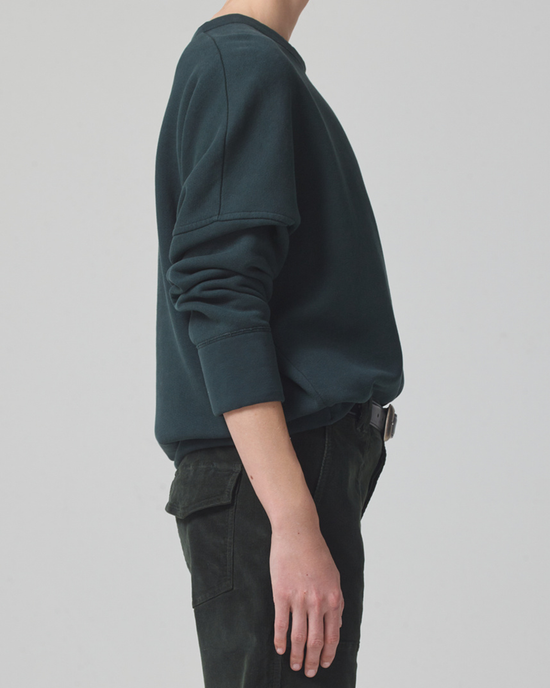A person standing sideways wearing an oversized dark green Citizens of Humanity Sam sweatshirt in Subert and black pants.