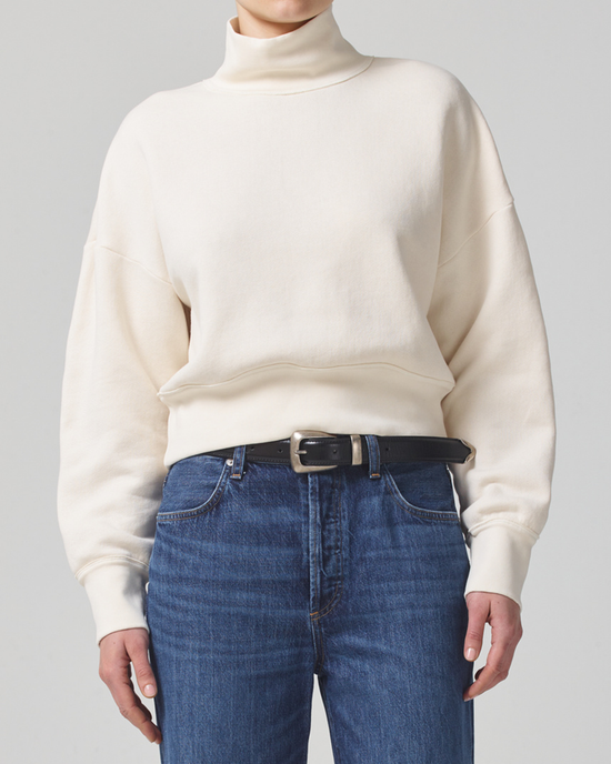A person wearing a cream Citizens of Humanity Koya Turtleneck in Canvas sweater and blue jeans with a black belt.