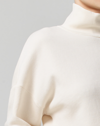 Close-up of a person wearing a cream Citizens of Humanity Koya Turtleneck sweater in Canvas against a gray background.