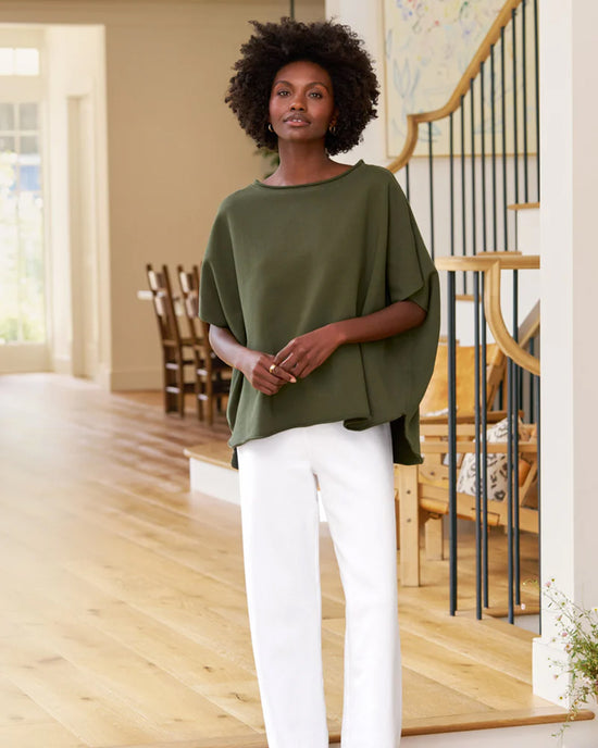 A woman with short curly hair poses confidently indoors beside a staircase, wearing Frank & Eileen's Olive Original Capelet in Caper and white pants in a modern, well-lit home with wooden floors.