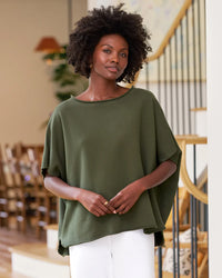 A woman with natural curly hair wears the Frank & Eileen Olive Original Capelet in Caper and white pants, standing indoors near a staircase with wooden rails and soft lighting.