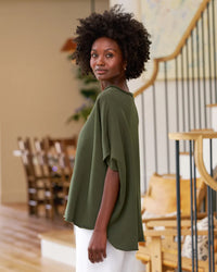 A woman with curly hair, indoors, wears the Frank & Eileen Olive Original Capelet in Caper and white pants, gazing at the camera. Cozy furniture and a wooden staircase are visible in the background.