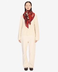 A person stands facing forward in a cream ribbed sweater, matching pants, black shoes, and an oversized Inoui Editions Square / Carre 130 Papillons scarf in vibrant red around their neck against a plain white background.