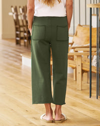 Indoors on a wooden floor, a person faces away in a white top and Frank & Eileen Penny Patch Pocket Sweatpant in Caper, featuring large back pockets, paired with white slide sandals. Wooden chairs and neutral décor appear in the background.