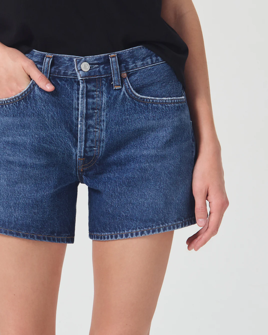 A person wearing AGOLDE's Parker Long Short in Enamour organic cotton denim shorts standing with their hand partially in the pocket.