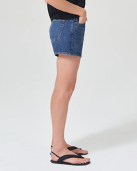 Woman wearing AGOLDE Parker Long Short in Enamour denim skirt and black flip-flops standing against a white background.