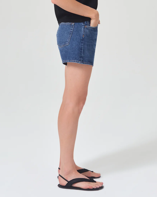 Woman wearing AGOLDE Parker Long Short in Enamour denim skirt and black flip-flops standing against a white background.