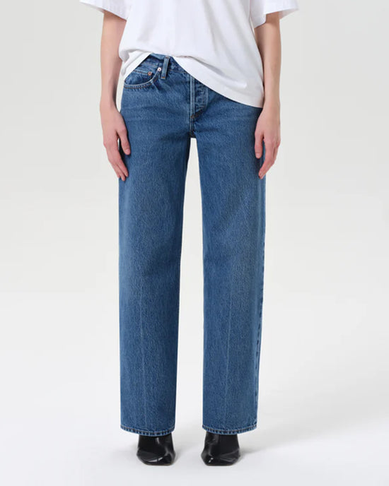 A person wearing a white T-shirt, AGOLDE Low Rise Loose in Charge denim jeans in blue, and black shoes stands against a plain light background.