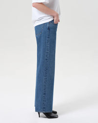 A person stands in profile against a plain light background wearing AGOLDE’s Low Rise Loose in Charge straight-leg blue jeans, regenerative cotton, paired with a white top and black heeled shoes.