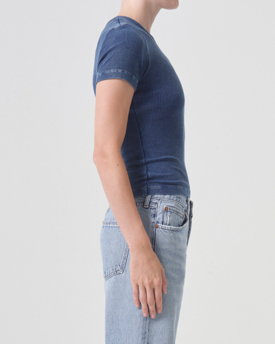 Side view of a woman wearing a ribbed cotton AGOLDE Arlo Rib Pocket Tee in Indigo and denim jeans against a light background.