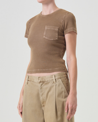 Woman wearing an AGOLDE Arlo Rib Pocket Tee in Bamboo with a chest pocket, paired with beige trousers, against a white background; only her torso and hips are visible.