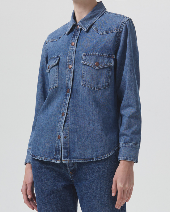 A person wearing a relaxed denim AGOLDE Glinda Shirt in Rhythm with front buttons and chest pockets stands against a plain background.