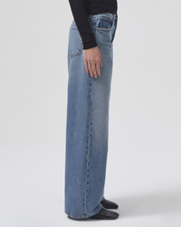 Person standing side-profile wearing Low Slung Baggy in Libertine jeans made of organic cotton denim by AGOLDE and black footwear.