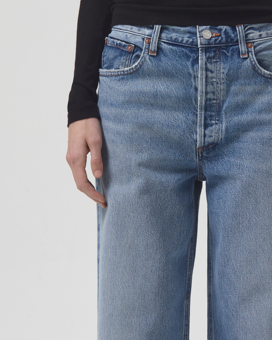 Close-up of a person wearing organic cotton denim AGOLDE Low Slung Baggy jeans in Libertine and a black top, with a focus on the denim texture and the person's hand resting along the seam.
