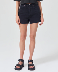 A person standing wearing black AGOLDE Dee Short in Slow Burn high rise denim shorts and black sandals.