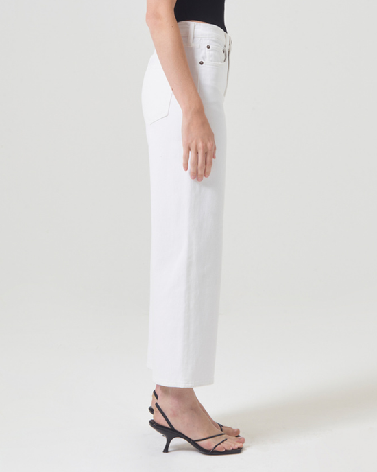 Side view of a person wearing white AGOLDE Harper Crop in Sour Cream jeans and black sandals standing against a plain background.