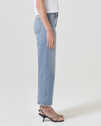 Side profile of a person wearing AGOLDE Harper Straight Crop in Hassle mid rise straight leg jeans and black strappy heels standing against a neutral background.