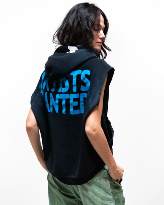 A woman in a Free City AW Cutoff Superyumm Biggie Hoodie in Superblack and green pants, viewed from the side, with the text "FREECITY artists wanted" in bold blue letters on the back of the hoodie.